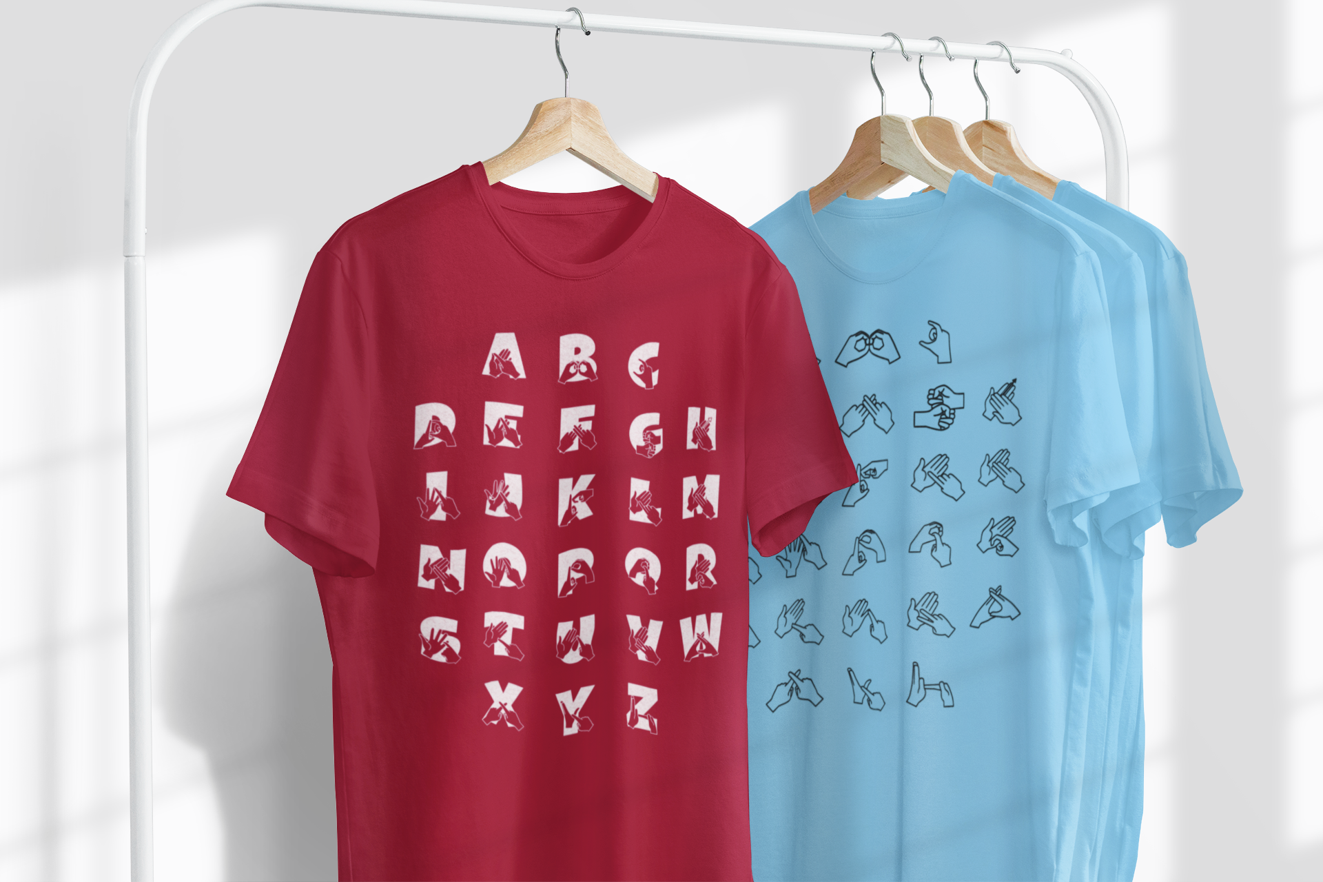 Four t-shirts on hangers. A red t-shirt shows the full BSL alphabet in white with English letters beneath. Next to it, a blue t-shirt displays the BSL alphabet in black fingerspelling only. Two more blue t-shirts are partially visible.