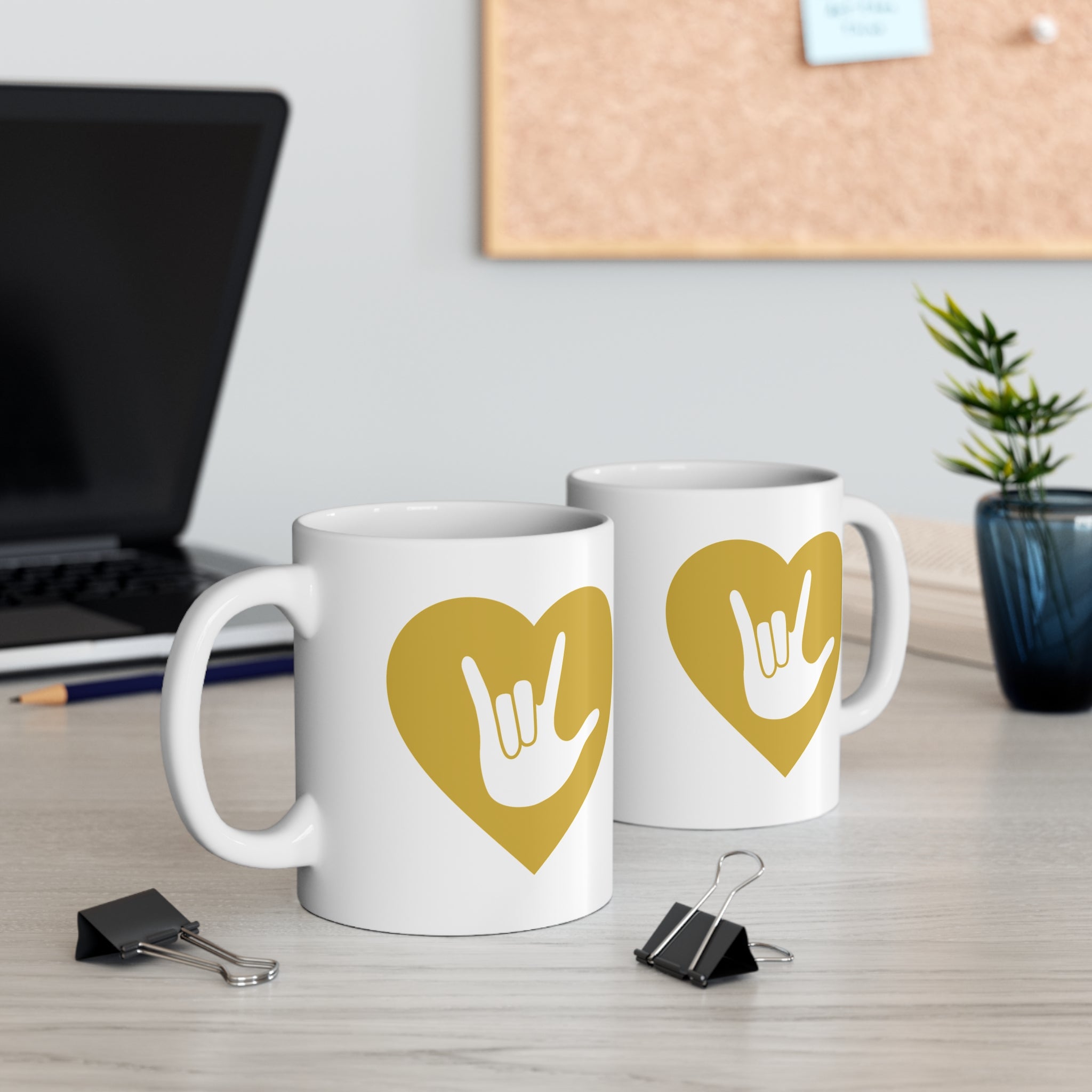Two mugs with gold coloured heart designs featuring the ASL sign for I Love You embedded in them on a desk with office supplies.