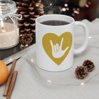 White mug with gold coloured heart design featuring the ASL sign for I Love You embedded in it on a table with decorative items.