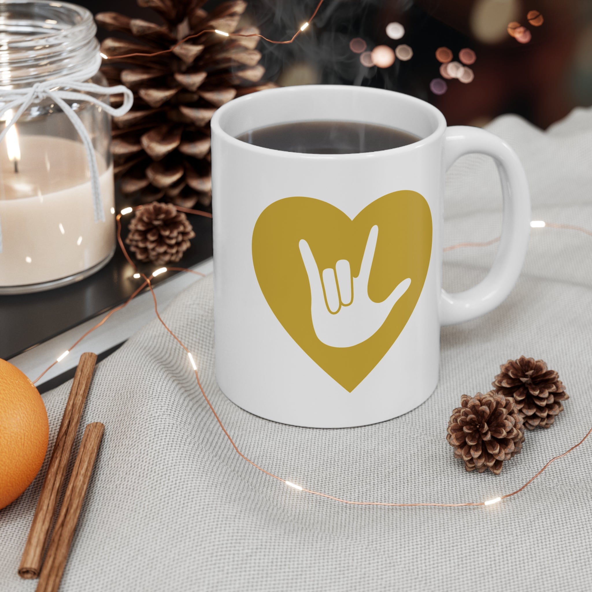 White mug with gold coloured heart design featuring the ASL sign for I Love You embedded in it on a table with decorative items.