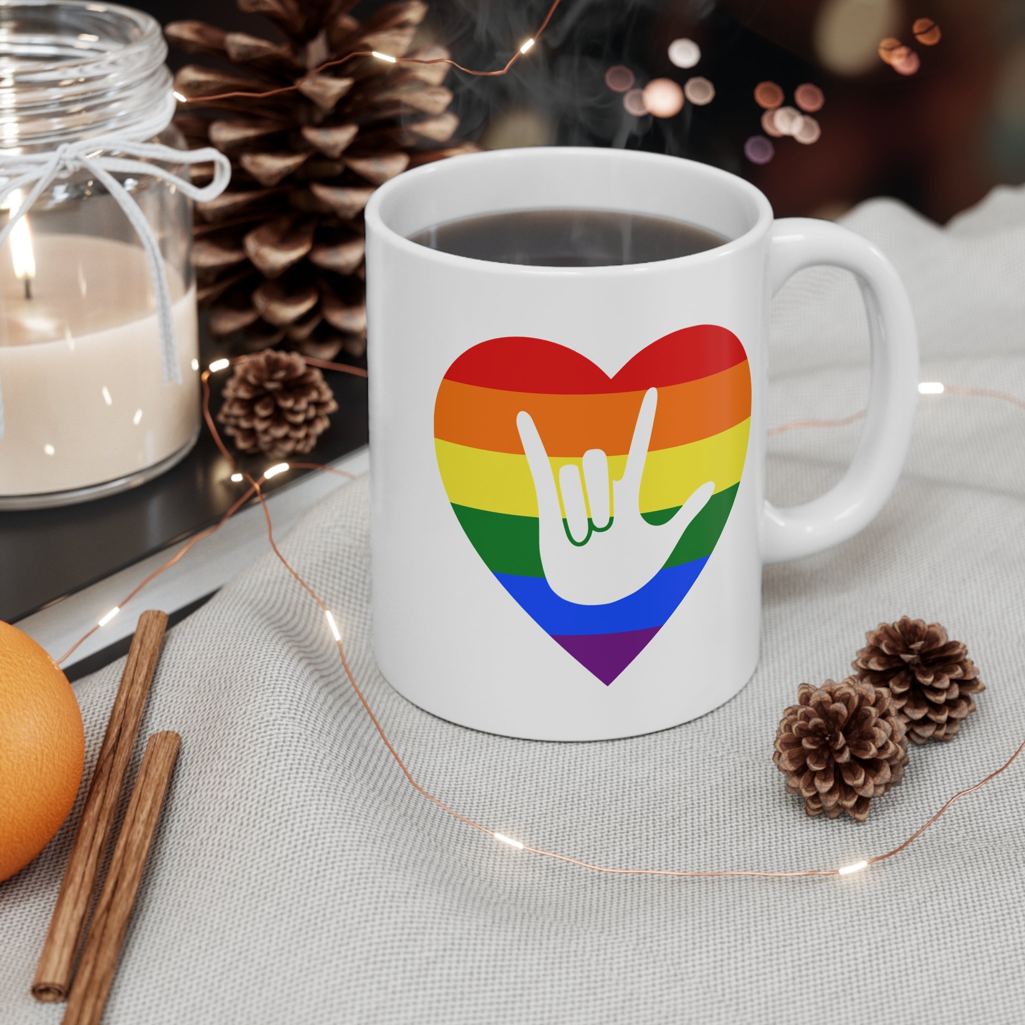 White mug with pride coloured heart design featuring the ASL sign for I Love You embedded in it on a table with decorative items