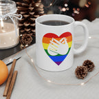 White mug with pride coloured heart design featuring the BSL sign for Love embedded in it on a table with decorative items.