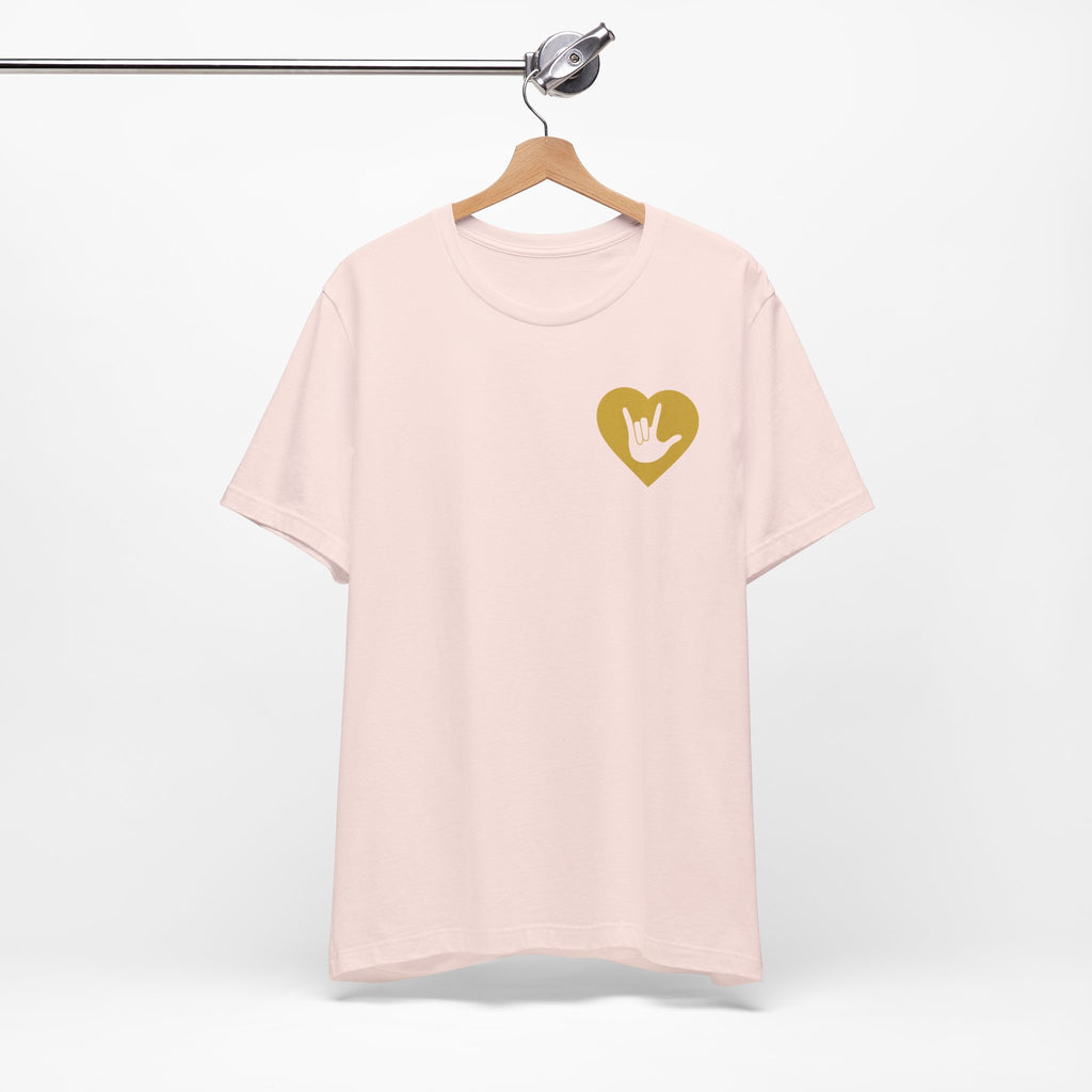 Front view of a soft pink T-Shirt showing a gold coloured heart and the iconic ASL I Love You sign embedded in it on the left side of the chest, on a hanger against a white background