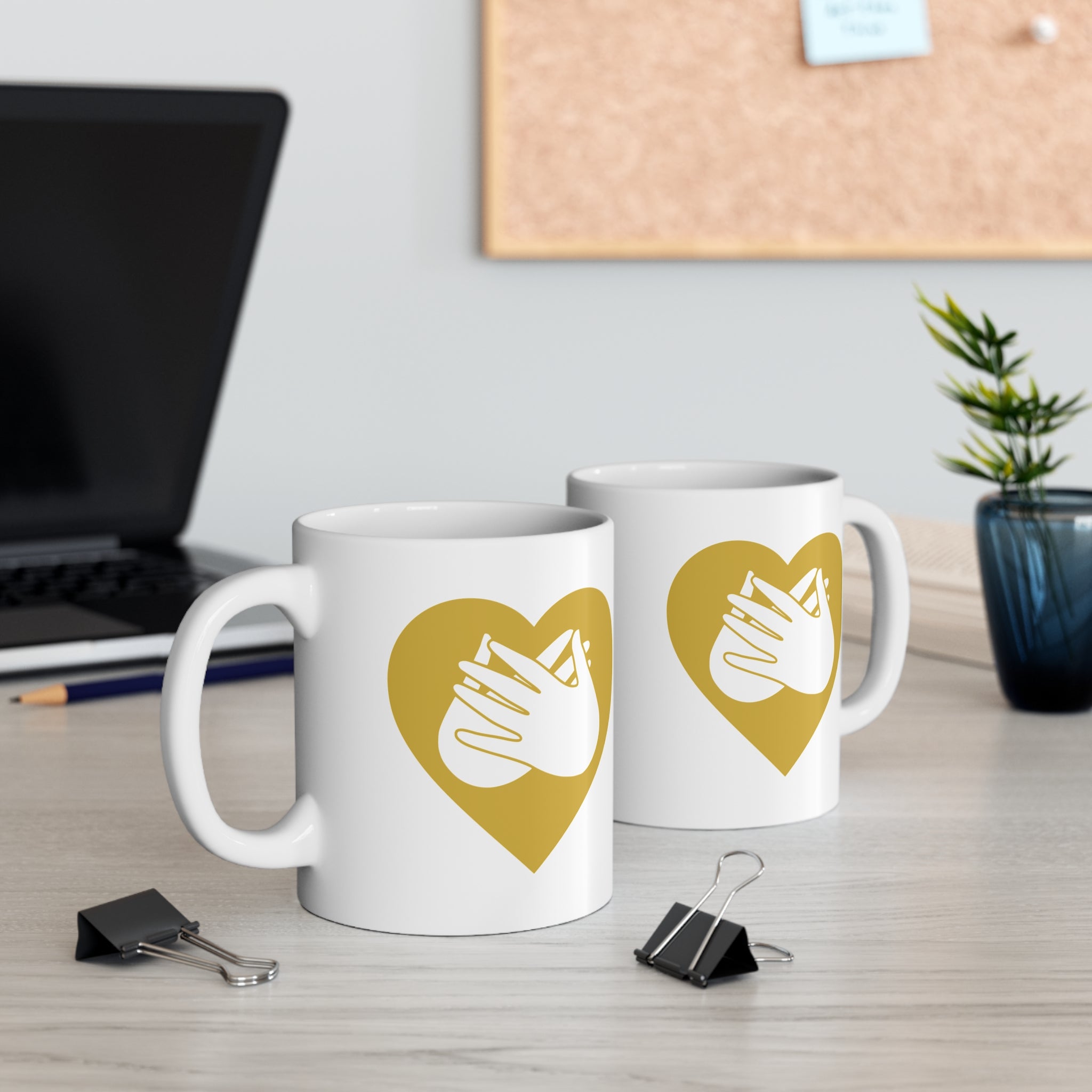 Two mugs with gold coloured heart designs featuring the BSL sign for Love embedded in them on a desk with office supplies.