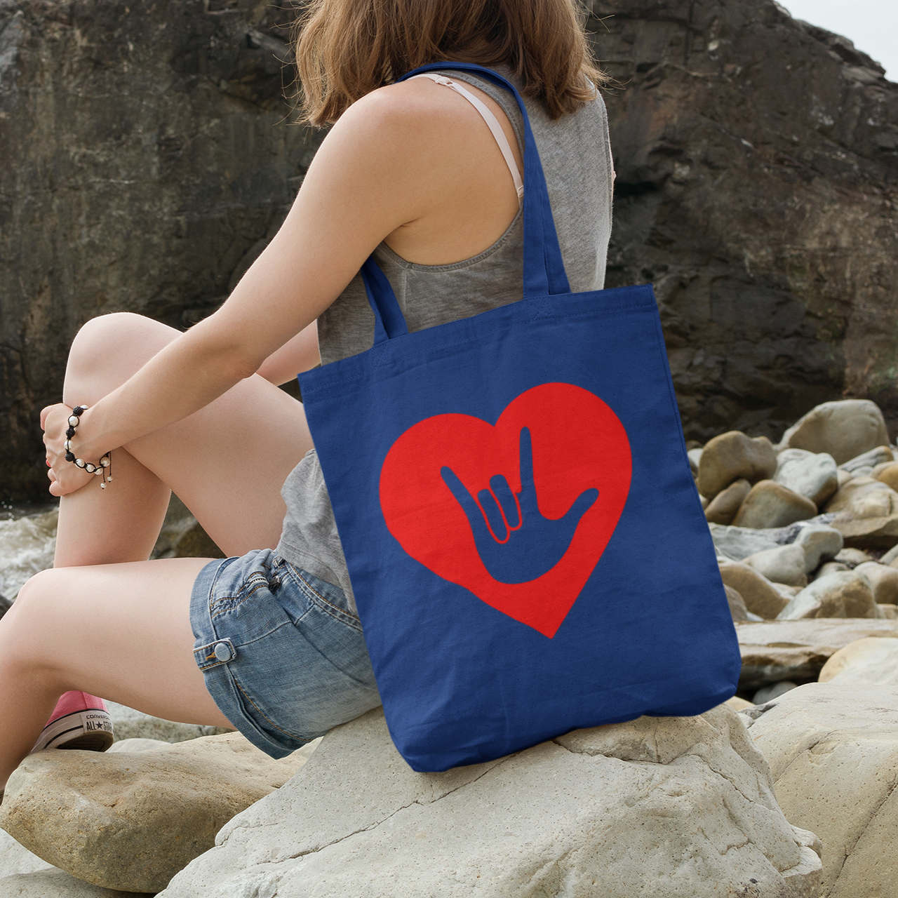 Person sitting on rocks by the water with a royal blue Tote Bag showing a gold coloured heart with the iconic ASL I Love You sign embedded in it