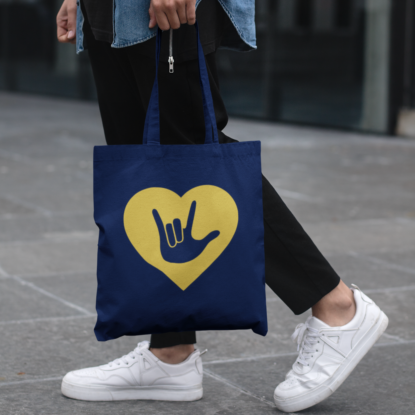 Person walking on a pavement holding a navy blue Tote Bag showing a gold coloured heart with the iconic ASL I Love You sign embedded in it.