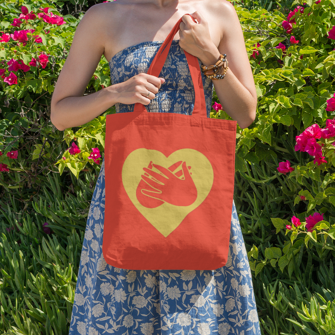 Person holding a Red Tote Bag showing a gold coloured heart and the iconic BSL Love sign embedded in it, stood in front of pink flowers.