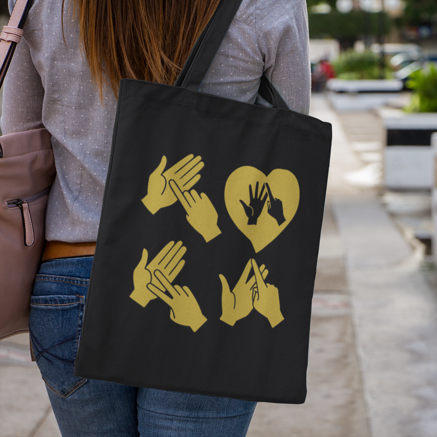 Person holding a black Tote Bag with the word Love in gold BSL Fingerspelling with the 'O' embedded in a heart shape design outdoors.