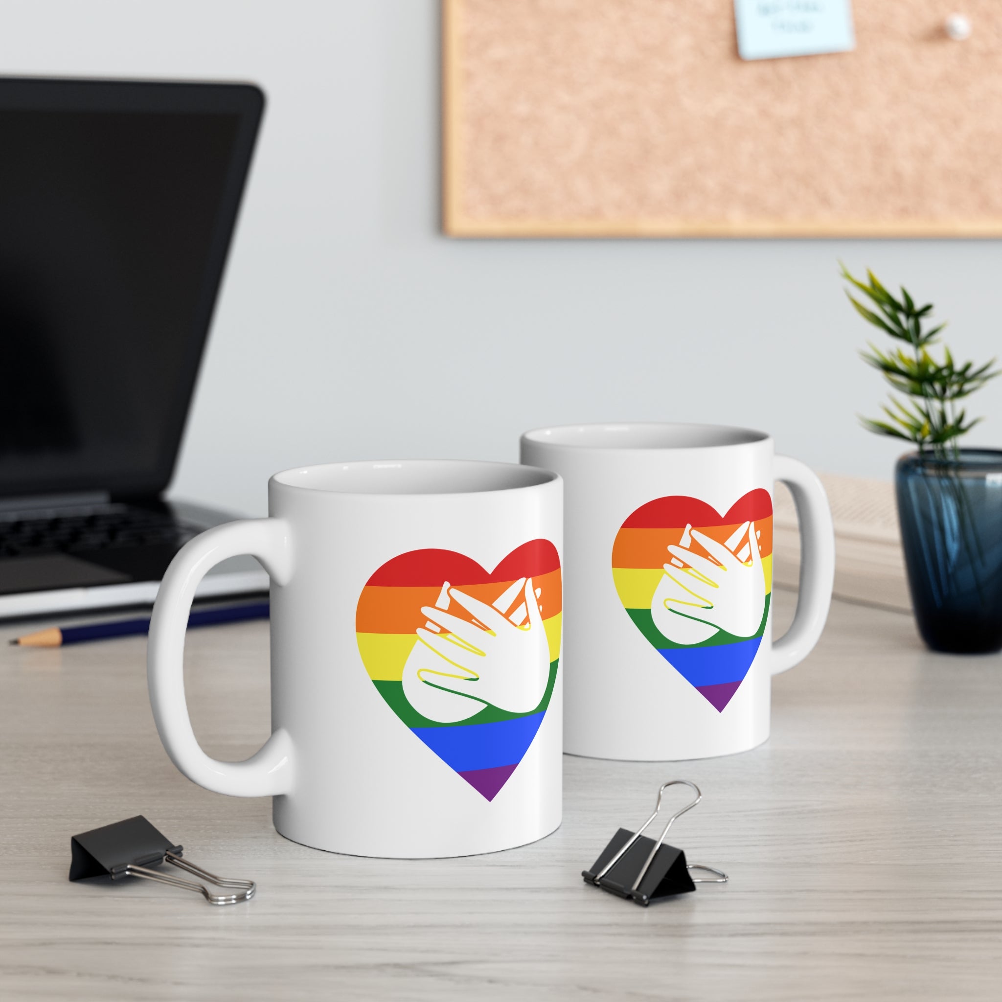 Two mugs with pride coloured heart designs featuring the BSL sign for Love embedded in them on a desk with office supplies.