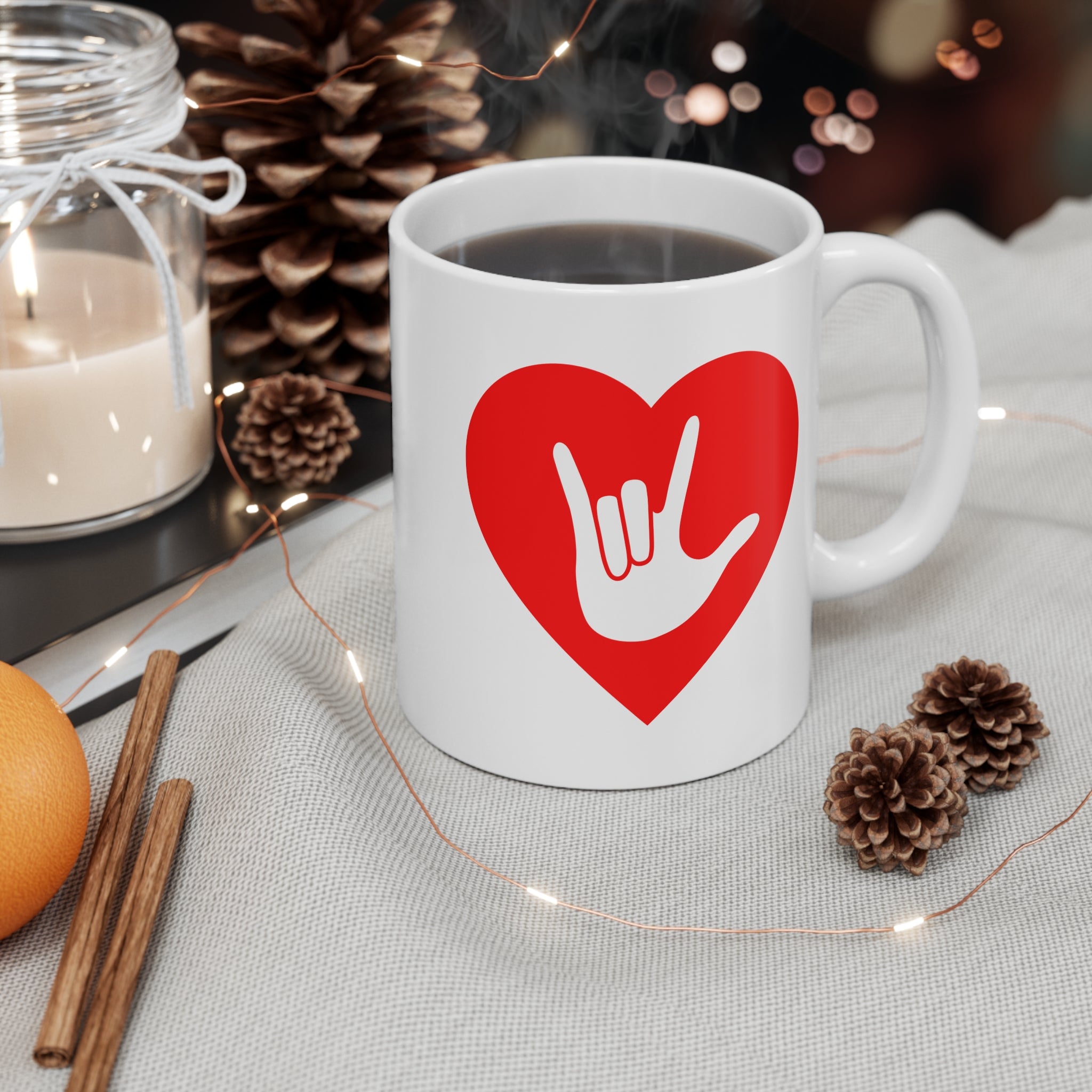 White mug with red coloured heart design featuring the ASL sign for I Love You embedded in it on a table with decorative items.