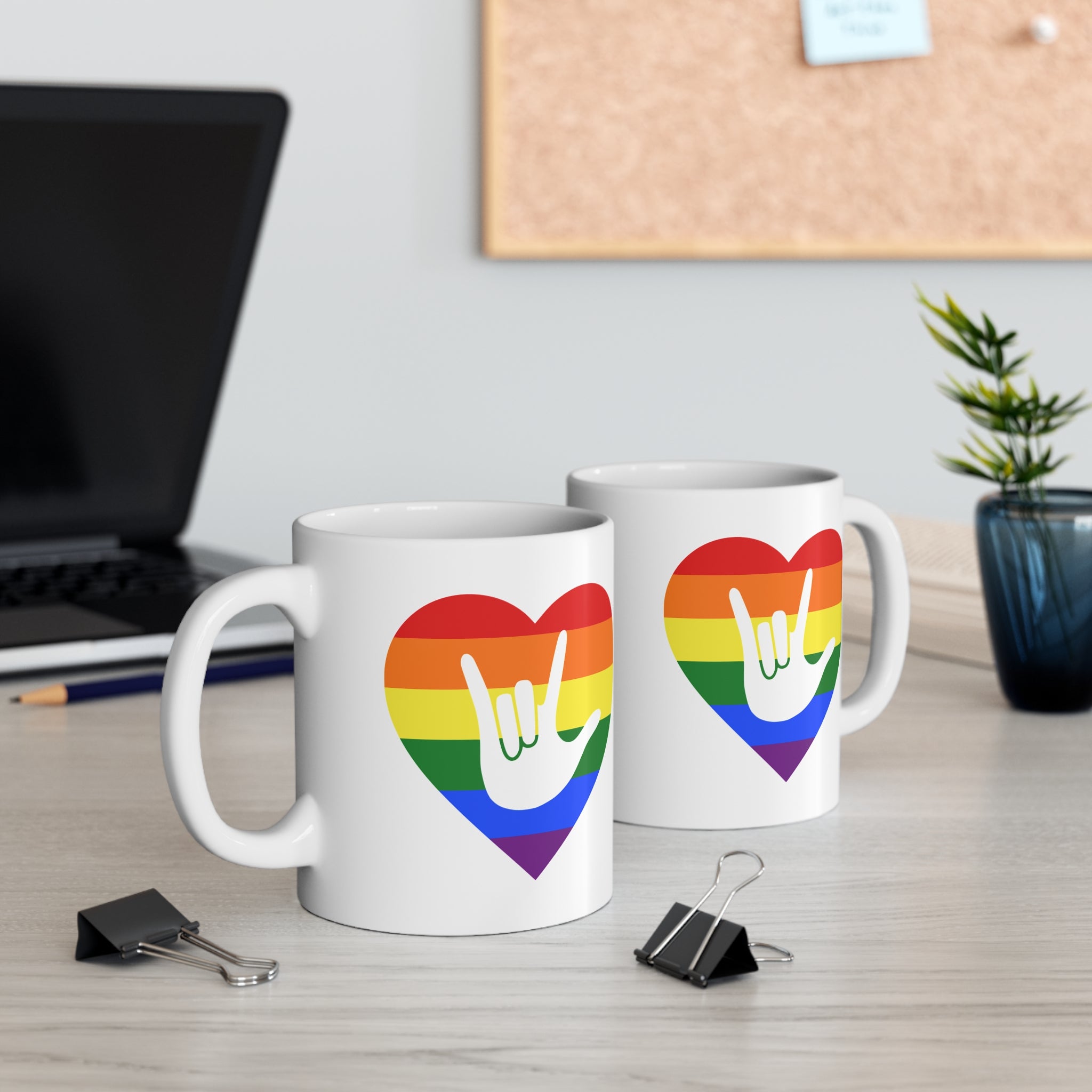 Two mugs with pride coloured heart designs featuring the ASL sign for I Love You embedded in them on a desk with office supplies.