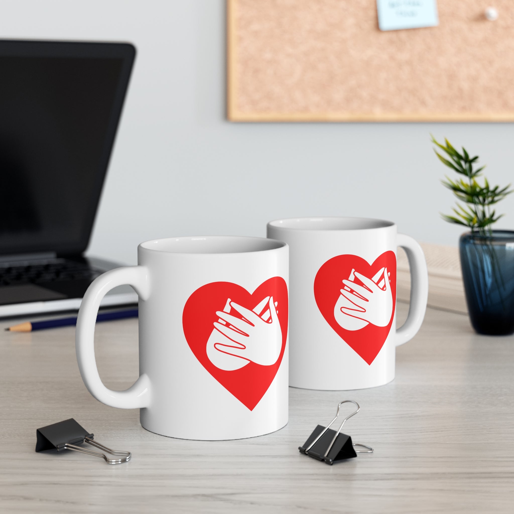 Two mugs with red coloured heart designs featuring the BSL sign for Love embedded in them on a desk with office supplies.