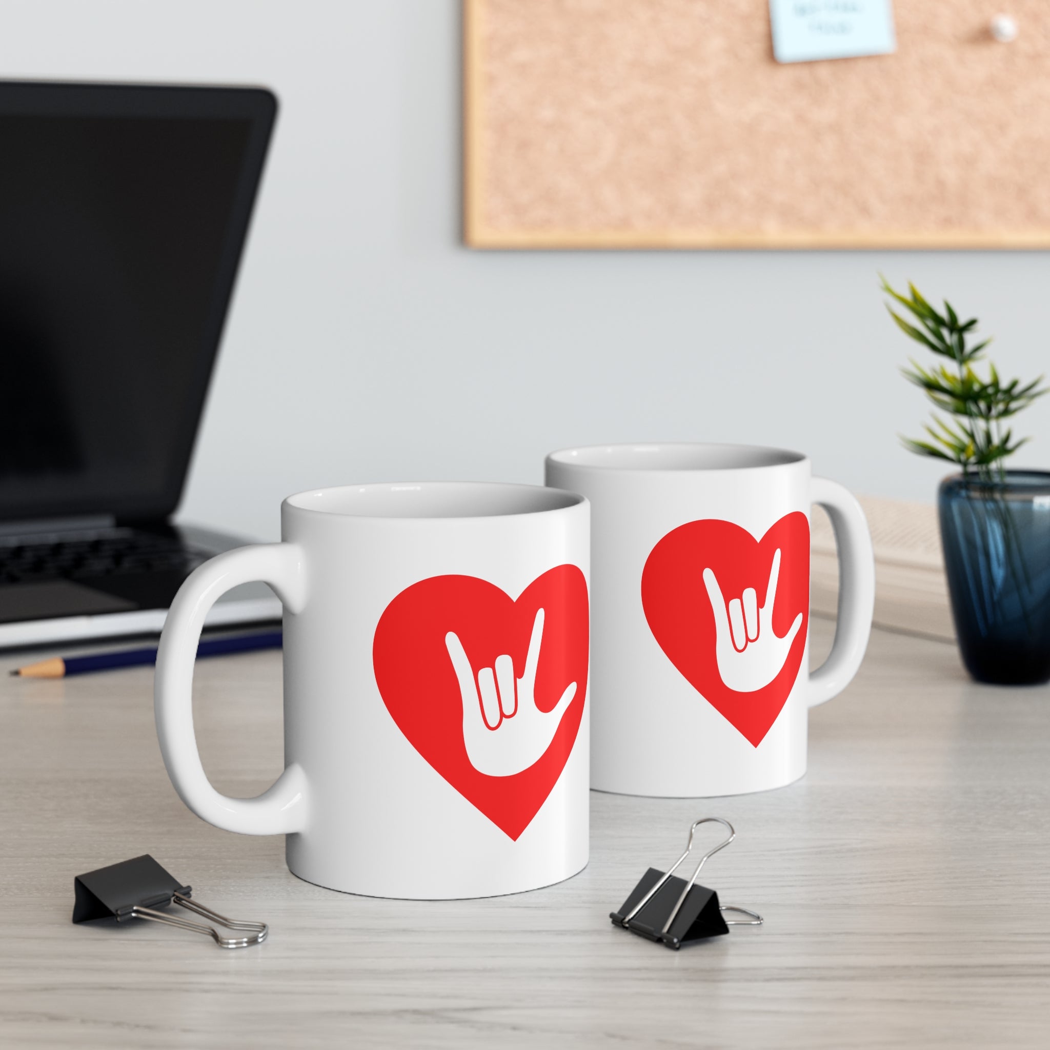 Two mugs with red coloured heart designs featuring the ASL sign for I Love You embedded in them on a desk with office supplies.