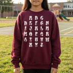 Child wearing a maroon sweatshirt with white alphabet print on a grassy area with playground equipment in the background.