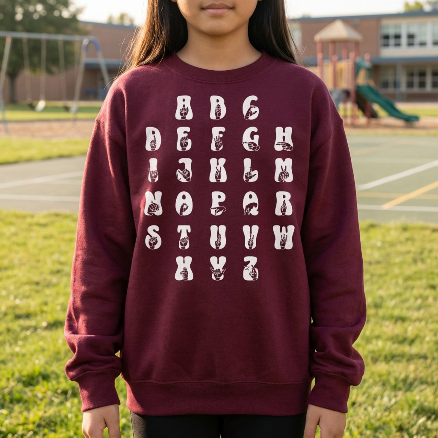 Child wearing a maroon sweatshirt with white alphabet print on a grassy area with playground equipment in the background.