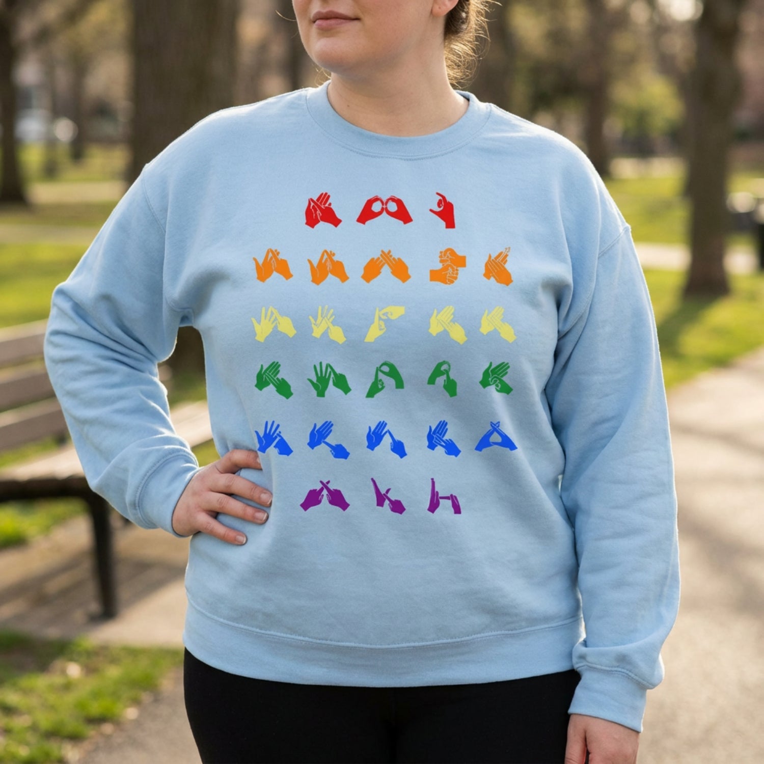 Person wearing a light blue sweatshirt with pride rainbow coloured fingerspelling in a park setting