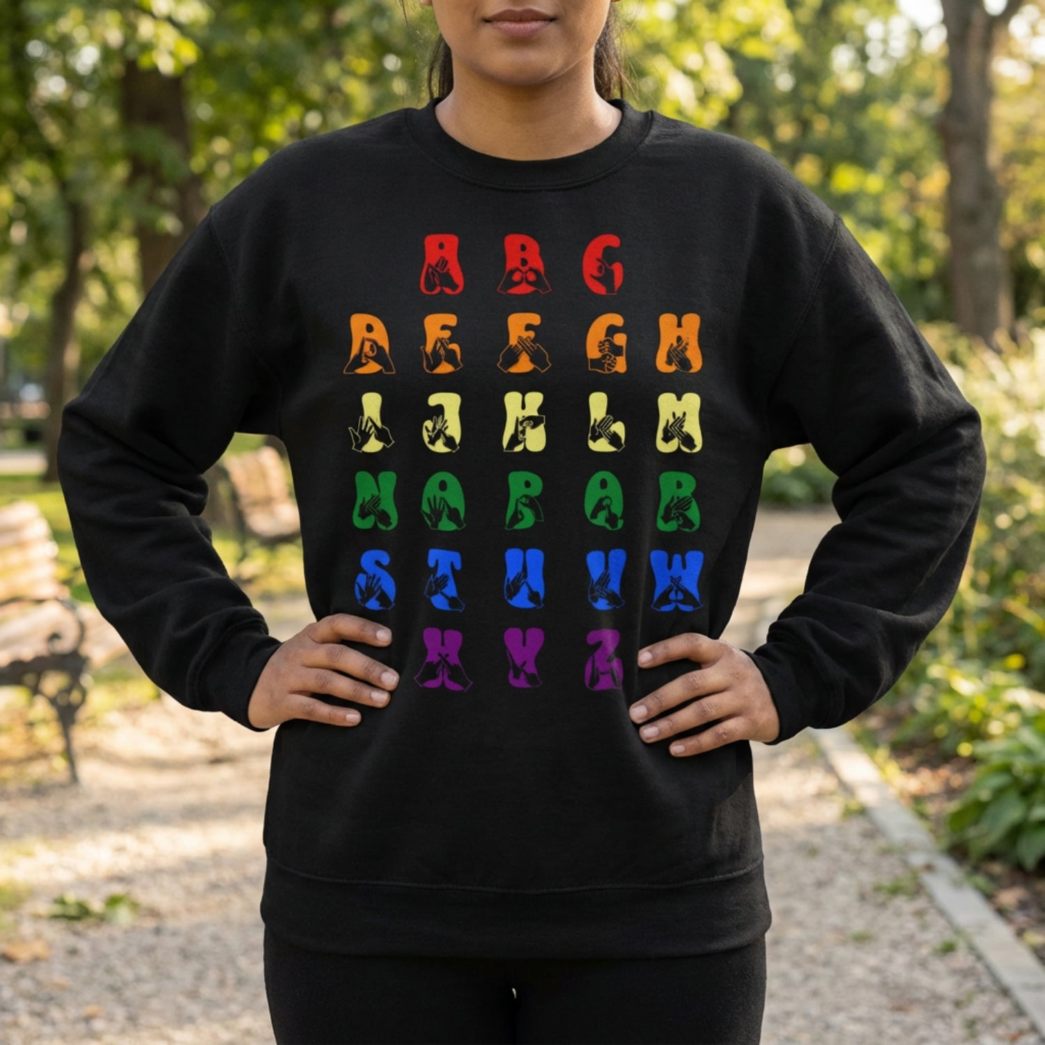 Person wearing a black sweatshirt with pride rainbow coloured alphabet design in a park setting