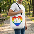 Person holding a tote bag with a rainbow heart design featuring two hands in the center, set against a park background.