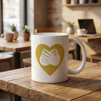 White mug with a gold heart and hand symbol on a wooden table in a cozy cafe setting.