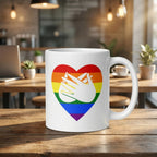 White mug with a pride rainbow heart and hand symbol on a wooden table in a cozy cafe setting.