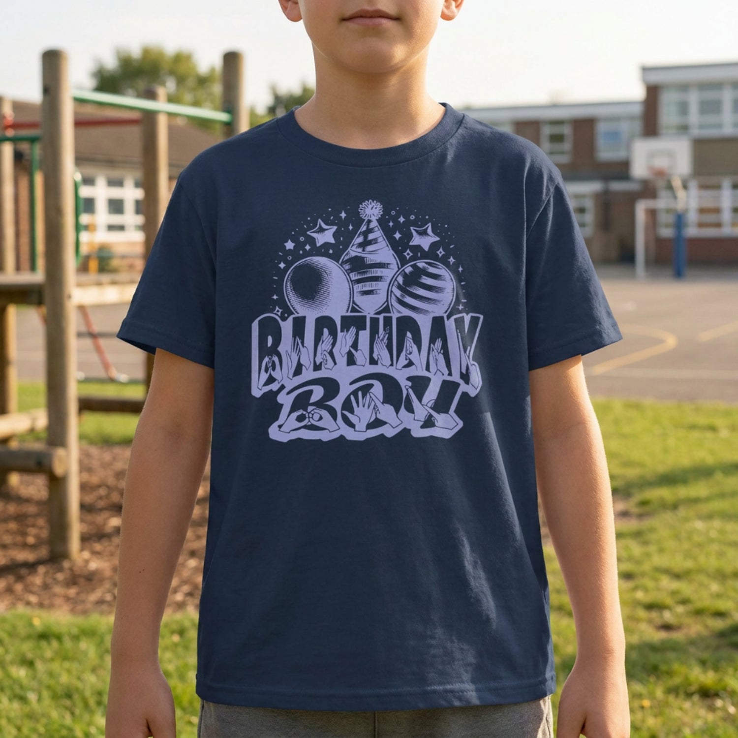 A person wearing a navy kids' t-shirt with "Birthday Boy" in bold letters featuring British Sign Language fingerspelling, with blue balloons, stars, and a party hat above.