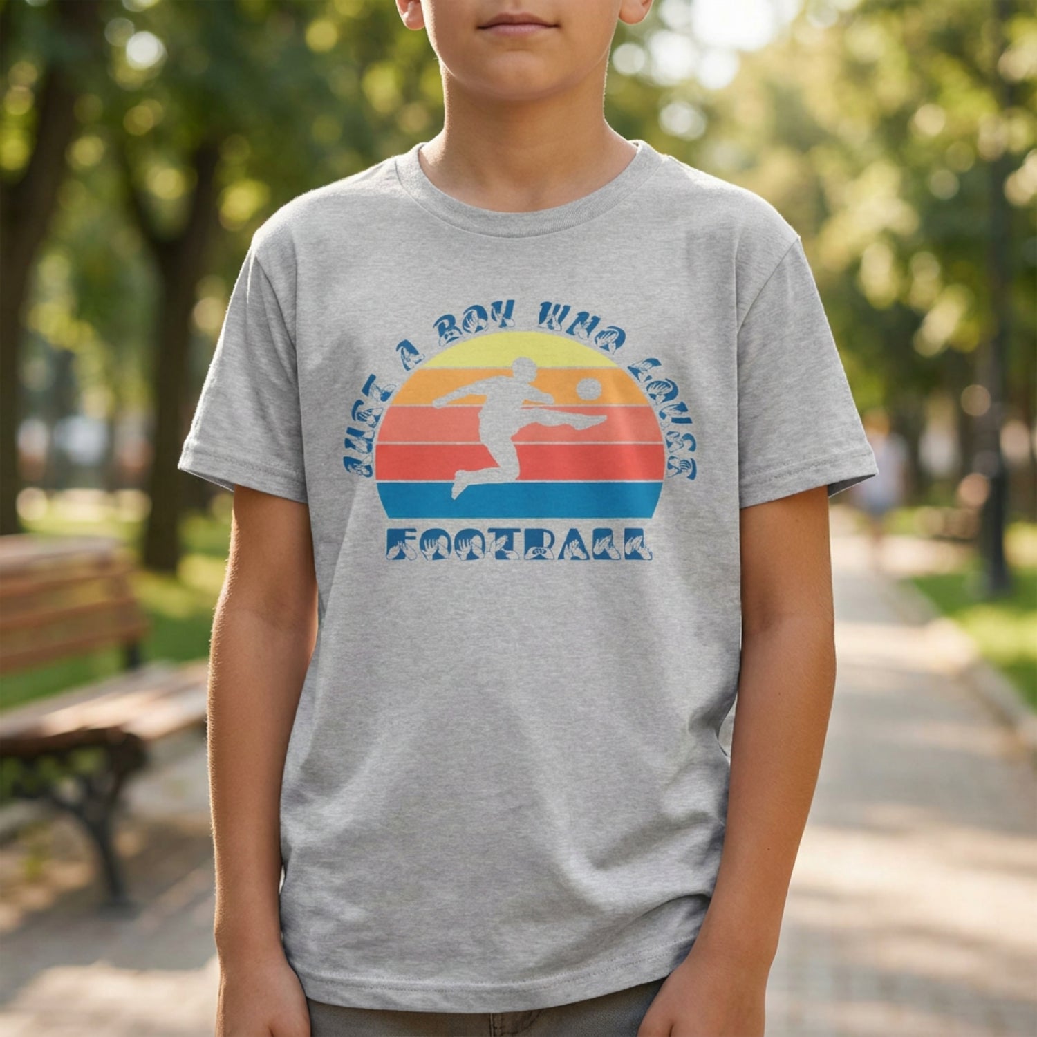 A boy wearing a sport grey t-shirt with the phrase "Just a Boy Who Loves Football" in BSL Fingerspelling and text featuring a colourful retro sunset and a silhouette of a boy kicking a football.