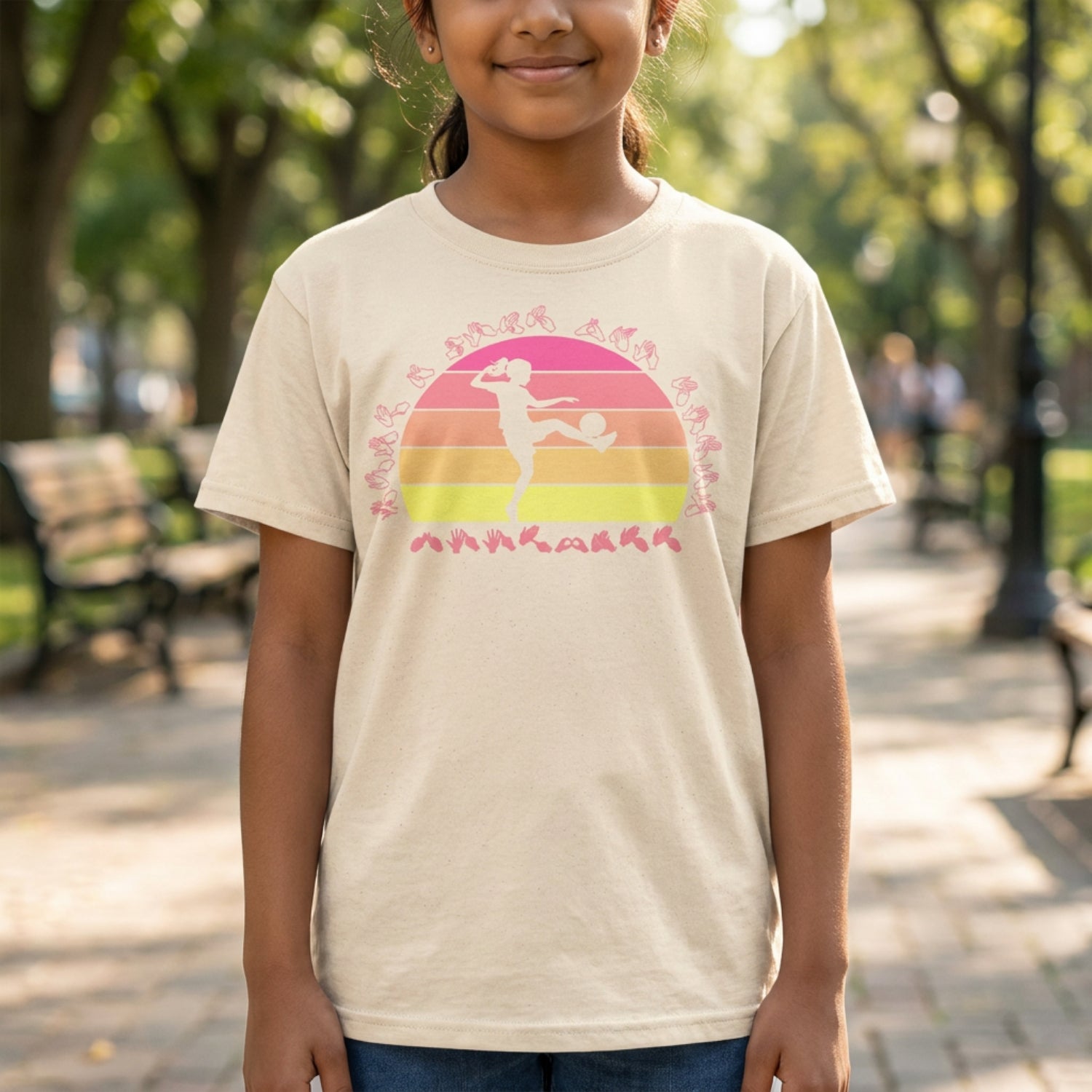 A person wearing a natural t-shirt with the phrase "Just a Girl Who Loves Football" in BSL Fingerspelling featuring a colourful retro sunset and a silhouette of a girl kicking a football.