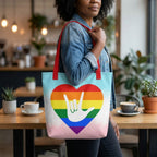 Person holding a tote bag with a rainbow heart and hand design in a cafe setting