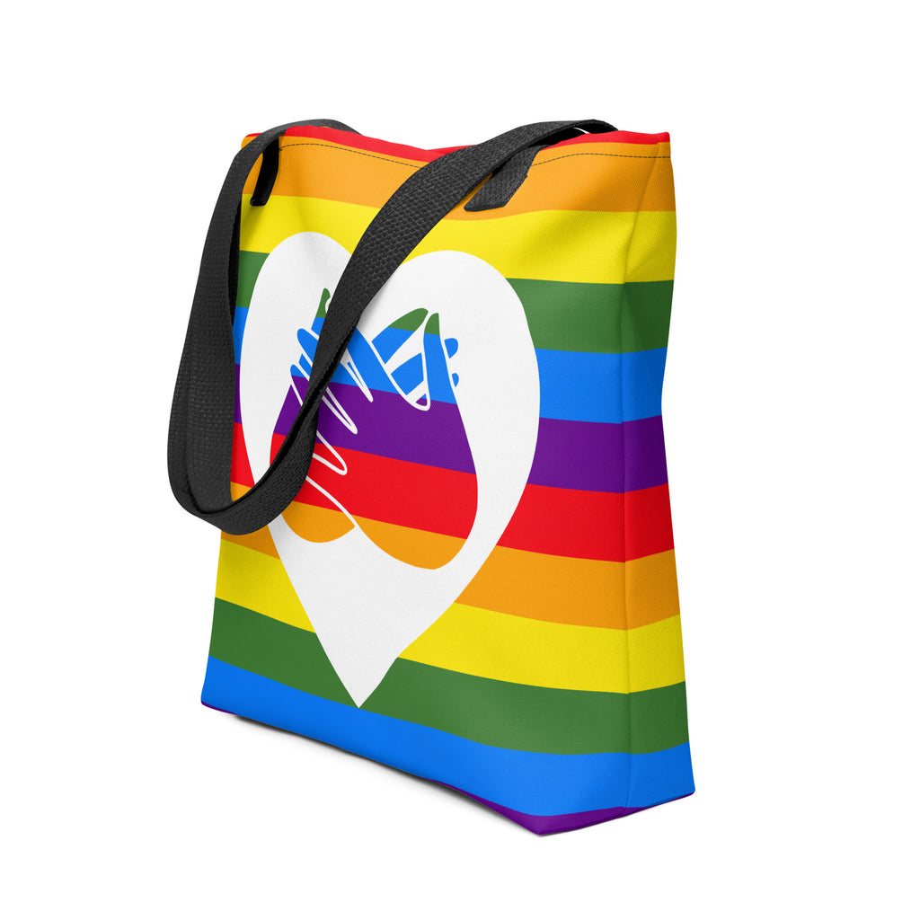 An angled view of a pride rainbow coloured tote bag with a black handle and a white heart with the BSL love sign at its centre.