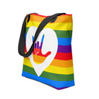 An angled view of a pride rainbow coloured tote bag with a black handle and a white heart with the ASL I love you sign at its centre.