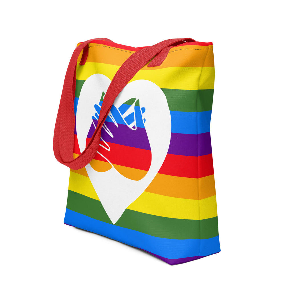 An angled view of a pride rainbow coloured tote bag with a red handle and a white heart with the BSL love sign at its centre.
