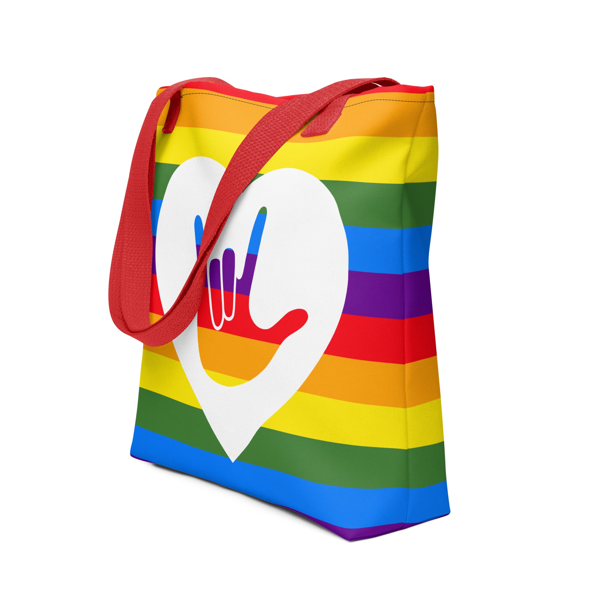 An angled view of a pride rainbow coloured tote bag with a red handle and a white heart with the ASL I love you sign at its centre.